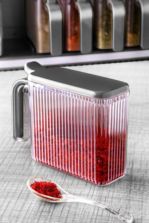 Gros plan sur un bocal à épices transparent avec poignée et couvercle gris, rempli d'épices rouges, accompagné d'une cuillère doseuse transparente.